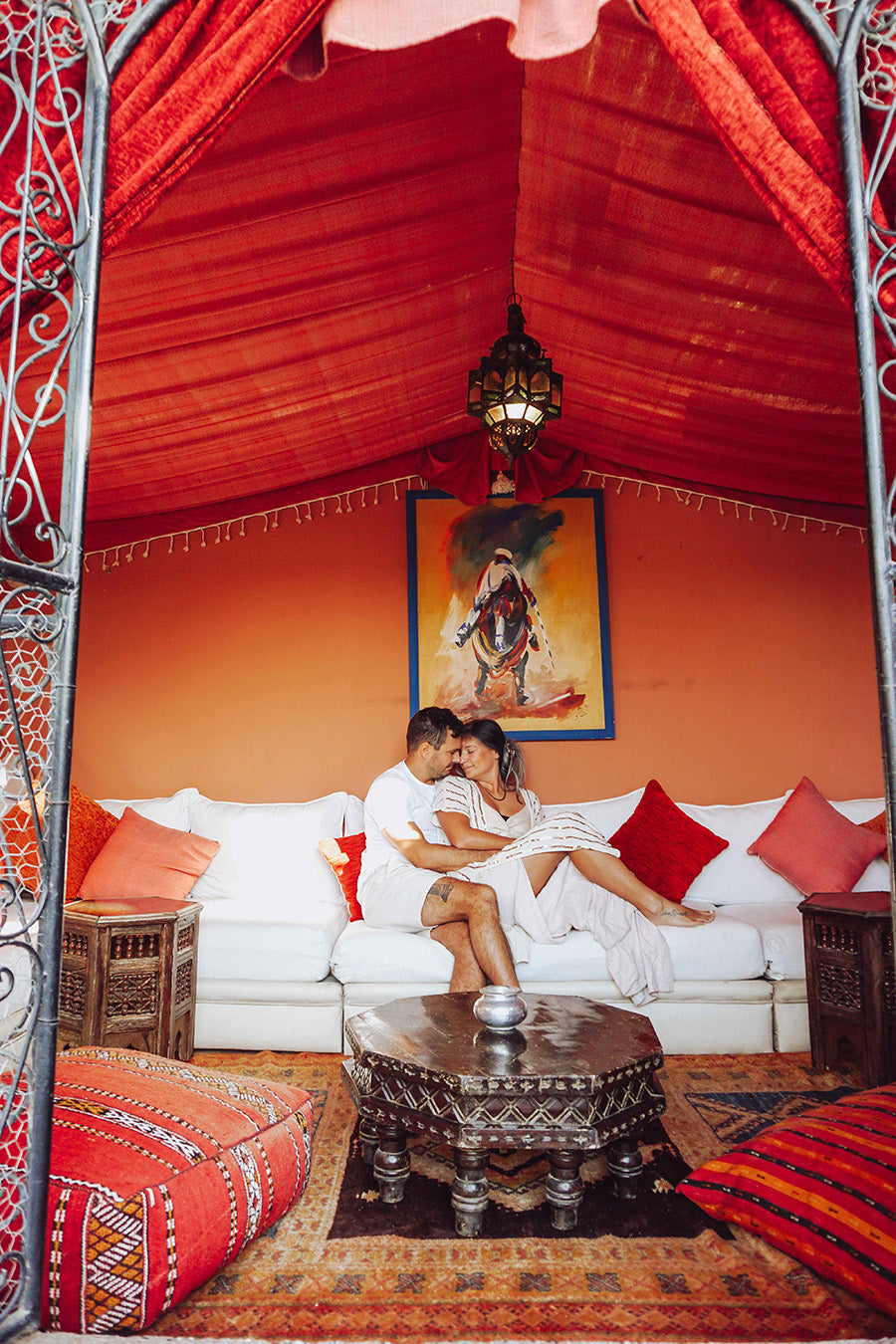 Couple embracing on a white sofa in a vibrant room with red and orange walls, decorative pillows, and a painting.